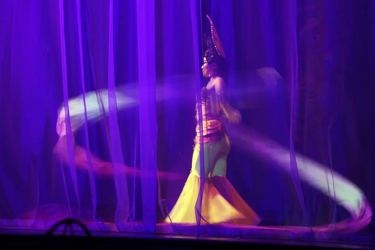 A dancer in a tall headdress and long skirt swirls pale silk ribbons around her on a stage lit in violet, seen through sheer curtains