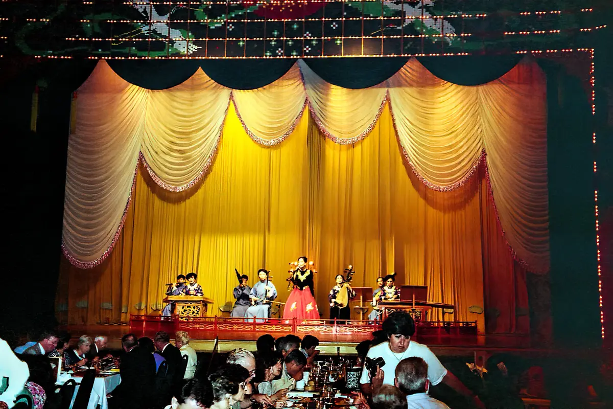 Diners at banquet tables in a dark hall face a stage hung with gold curtains, where a singer in red stands among seated musicians playing Chinese instruments